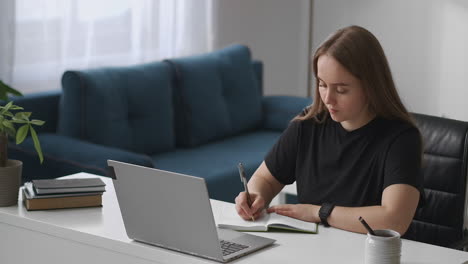 young-student-woman-is-listening-to-lecture-by-video-in-laptop-and-writing-notes-in-notebook-distant-education-and-e-learning-medium-portrait-of-female-in-living-room